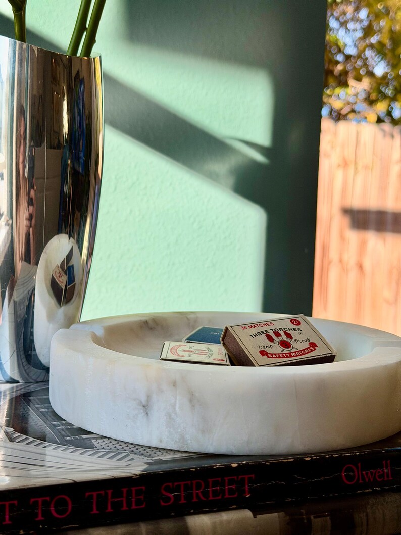 Puede incluir: Un cenicero de m&aacute;rmol blanco con un dise&ntilde;o circular, que contiene dos tarjetas peque&ntilde;as y una caja de cerillas. La caja de cerillas dice "THREE TORCHES" y "SAFETY MATCHES". Tambi&eacute;n se ven un jarr&oacute;n plateado y un libro.