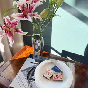 Puede incluir: Un bodeg&oacute;n con lirios rosas y blancos en un jarr&oacute;n plateado, libros y un cuenco de m&aacute;rmol blanco con cajas de cerillas. La escena est&aacute; sobre una mesa de madera, con sombras.