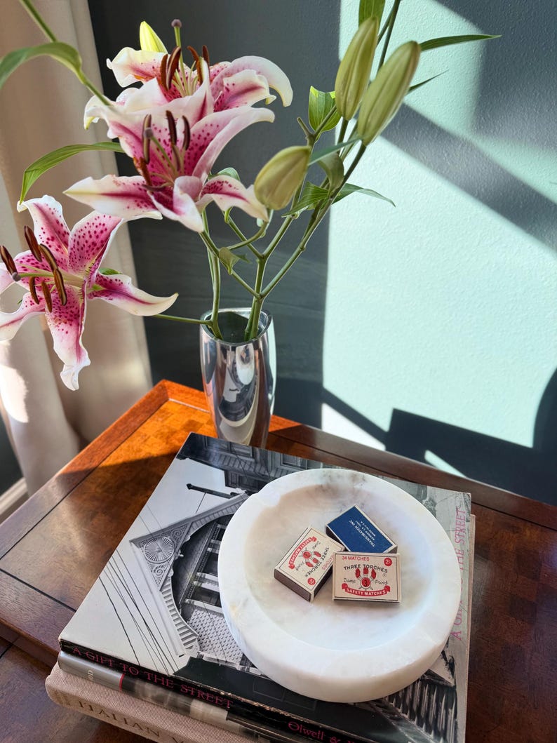 Puede incluir: Un bodeg&oacute;n con lirios rosas y blancos en un jarr&oacute;n plateado, libros y un cuenco de m&aacute;rmol blanco con cajas de cerillas. La escena est&aacute; sobre una mesa de madera, con sombras.