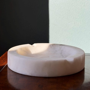 Puede incluir: Un cenicero redondo de m&aacute;rmol blanco. El cenicero tiene una superficie lisa y pulida y una hendidura curva para reposar cigarrillos. El cenicero es de color claro y est&aacute; sobre una superficie de madera oscura.