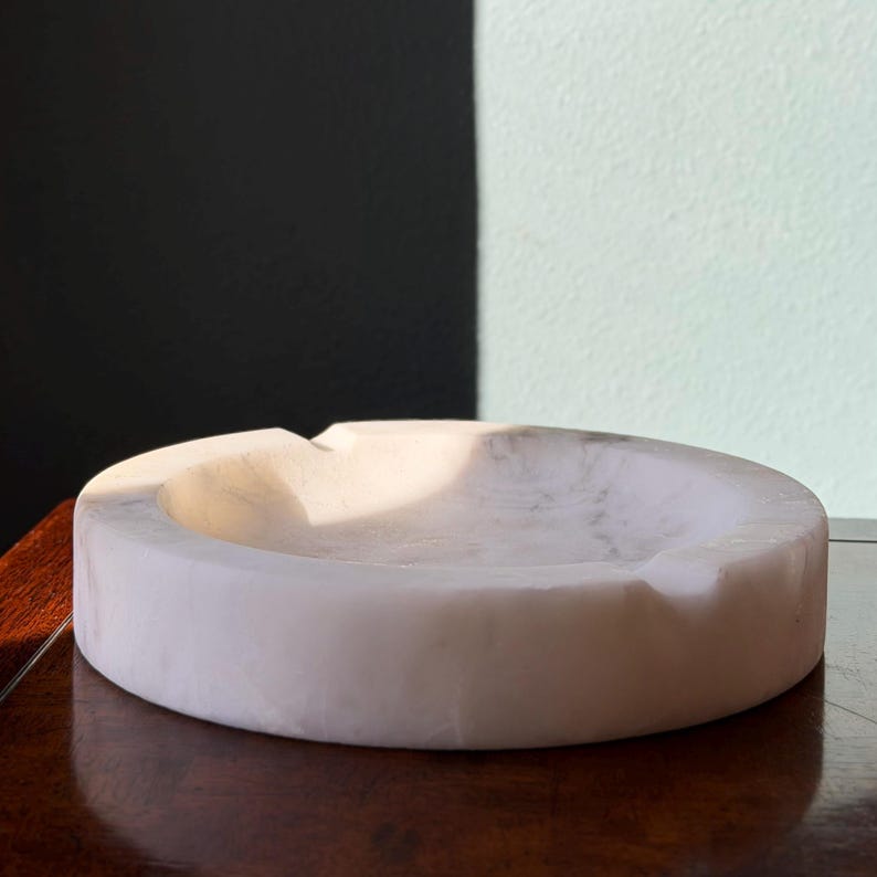 Puede incluir: Un cenicero redondo de m&aacute;rmol blanco. El cenicero tiene una superficie lisa y pulida y una hendidura curva para reposar cigarrillos. El cenicero es de color claro y est&aacute; sobre una superficie de madera oscura.