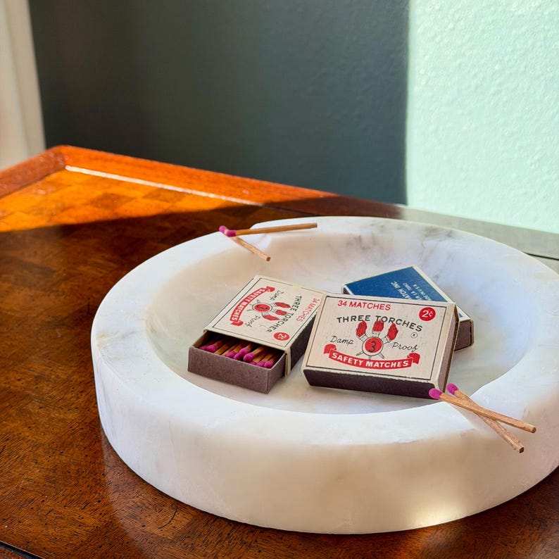 Puede incluir: Un cuenco de m&aacute;rmol blanco contiene dos cajas de cerillas abiertas y una caja de cerillas azul cerrada. Las cajas de cerillas son marrones con texto rojo y blanco. Se ven varias cerillas con puntas rosas.