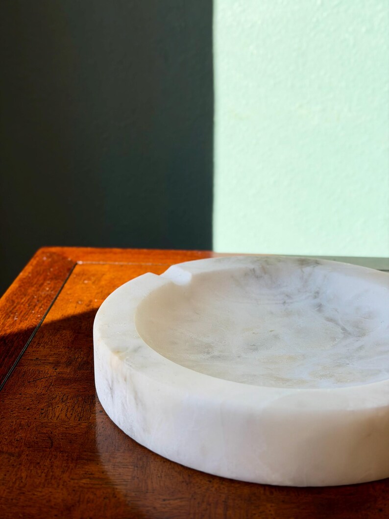 Puede incluir: Un cenicero de m&aacute;rmol blanco con forma redondeada y un solo reposacigarrillos. El cenicero est&aacute; sobre una superficie de madera oscura, con una pared verde oscuro y verde claro en el fondo. El m&aacute;rmol tiene un patr&oacute;n veteado natural.