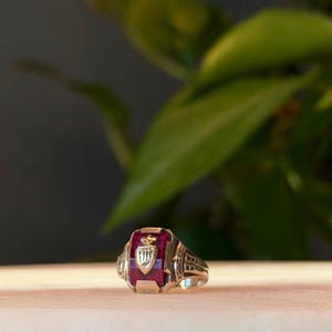 May include: A gold-colored class ring with a rectangular, ruby-red stone. The ring features intricate detailing on the sides and a crest design on the stone. The crest includes a shield and a crown.