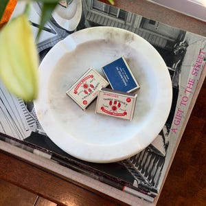 Puede incluir: Un cenicero redondo de m&aacute;rmol blanco con tres cajas de cerillas. Dos cajas son rojas y blancas, con "THREE TORCHES" y "SAFETY MATCHES" impresos. La tercera caja es azul con "TRANS/MATCH INC" impreso. El cenicero descansa sobre una pila de libros.