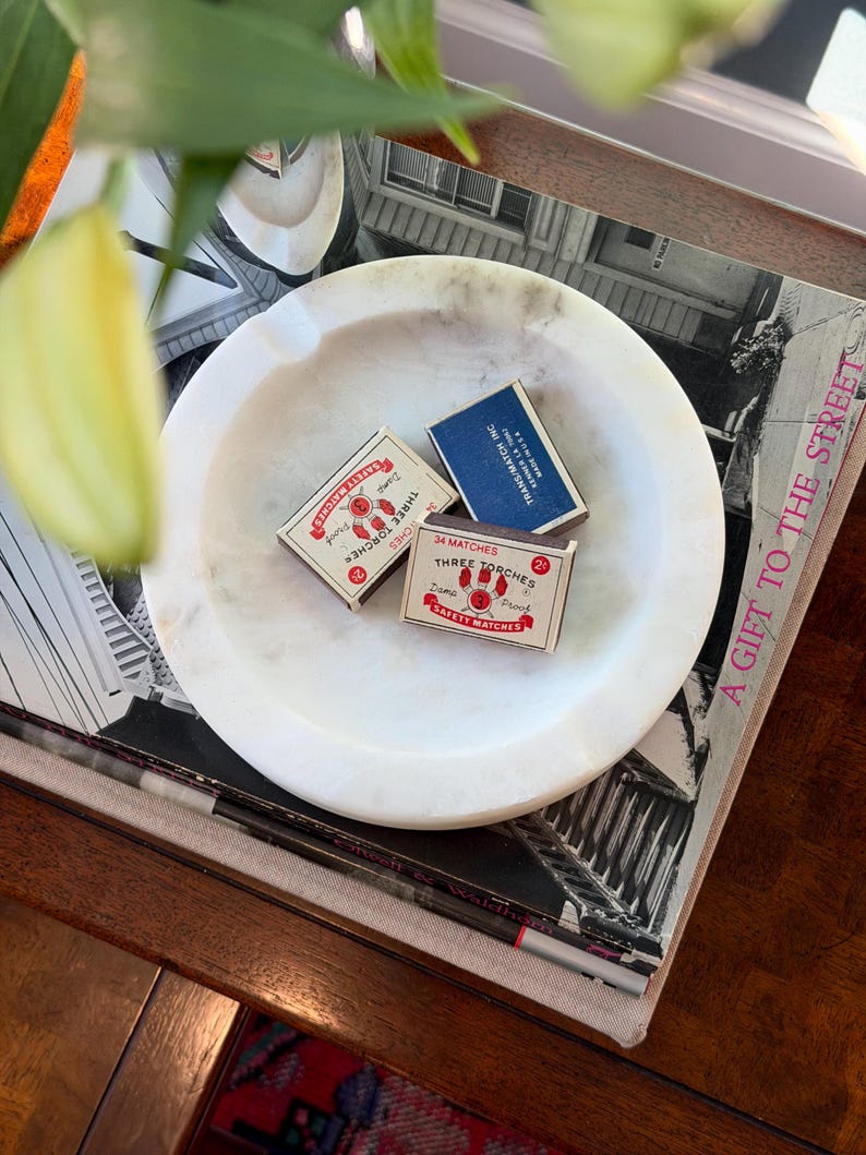 Puede incluir: Un cenicero redondo de m&aacute;rmol blanco con tres cajas de cerillas. Dos cajas son rojas y blancas, con "THREE TORCHES" y "SAFETY MATCHES" impresos. La tercera caja es azul con "TRANS/MATCH INC" impreso. El cenicero descansa sobre una pila de libros.