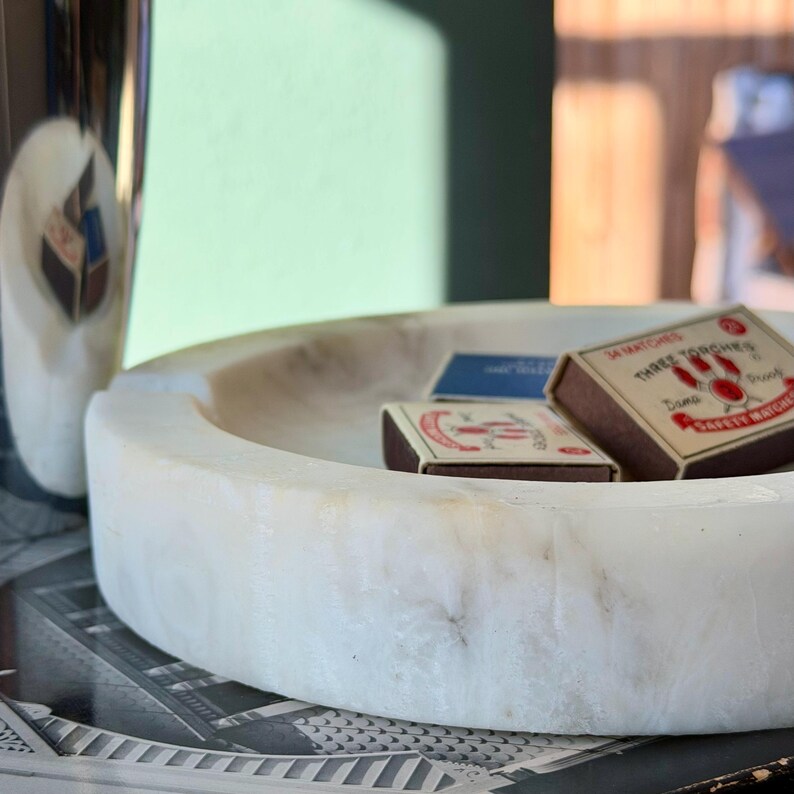 Puede incluir: Una bandeja redonda de m&aacute;rmol blanco contiene dos cajas de cerillas y una tarjeta azul. Las cajas de cerillas son marrones con texto rojo y blanco. Un jarr&oacute;n plateado y blanco es parcialmente visible a la izquierda. La bandeja descansa sobre una superficie impresa en blanco y negro.