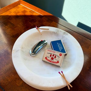 Puede incluir: Un cuenco de m&aacute;rmol blanco contiene un encendedor plateado, dos cajas de cerillas y varias cerillas con puntas moradas. Una caja de cerillas dice "34 MATCHES THREE TORCHES SAFETY MATCHES". El cuenco est&aacute; sobre una superficie de madera oscura.