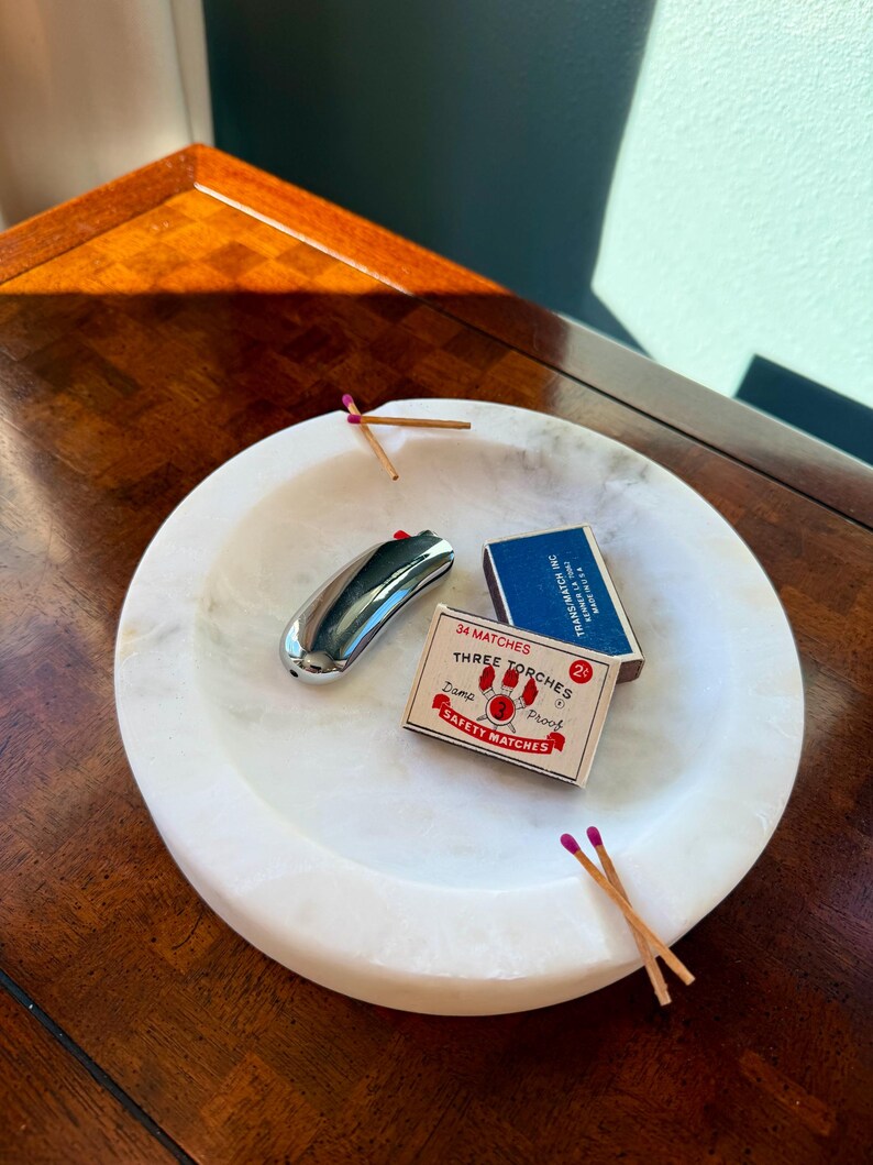 Puede incluir: Un cuenco de m&aacute;rmol blanco contiene un encendedor plateado, dos cajas de cerillas y varias cerillas con puntas moradas. Una caja de cerillas dice "34 MATCHES THREE TORCHES SAFETY MATCHES". El cuenco est&aacute; sobre una superficie de madera oscura.