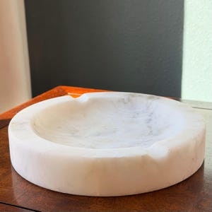 Puede incluir: Un cenicero redondo de m&aacute;rmol blanco con sutiles vetas grises. El cenicero tiene una superficie lisa y pulida y un dise&ntilde;o curvo con una peque&ntilde;a muesca para cigarrillos. Un art&iacute;culo decorativo para el hogar.