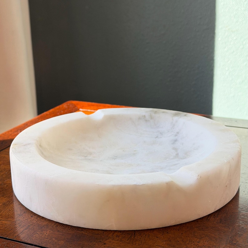 Puede incluir: Un cenicero redondo de m&aacute;rmol blanco con sutiles vetas grises. El cenicero tiene una superficie lisa y pulida y un dise&ntilde;o curvo con una peque&ntilde;a muesca para cigarrillos. Un art&iacute;culo decorativo para el hogar.