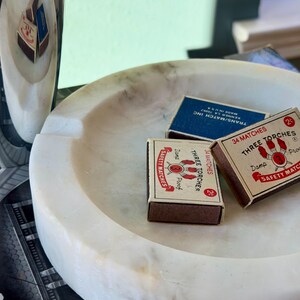 Puede incluir: Un cenicero de m&aacute;rmol blanco con tres cajas de cerillas. Dos cajas de cerillas son marrones con texto rojo y blanco, y una es azul con texto blanco. Un art&iacute;culo de m&aacute;rmol a juego est&aacute; en el fondo.