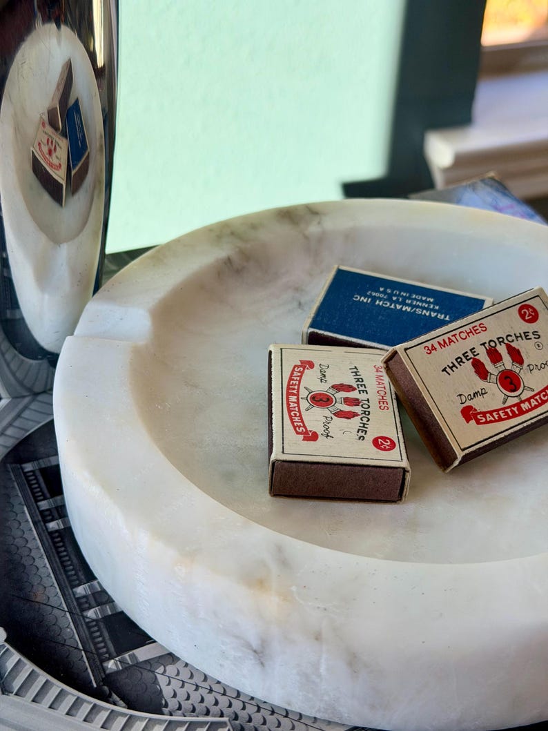 Puede incluir: Un cenicero de m&aacute;rmol blanco con tres cajas de cerillas. Dos cajas de cerillas son marrones con texto rojo y blanco, y una es azul con texto blanco. Un art&iacute;culo de m&aacute;rmol a juego est&aacute; en el fondo.
