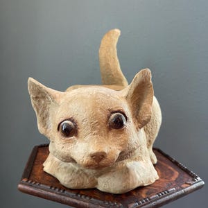 May include: A ceramic figurine of a tan-colored dog with large brown eyes. The dog is sitting on a wooden table with a hexagonal top.