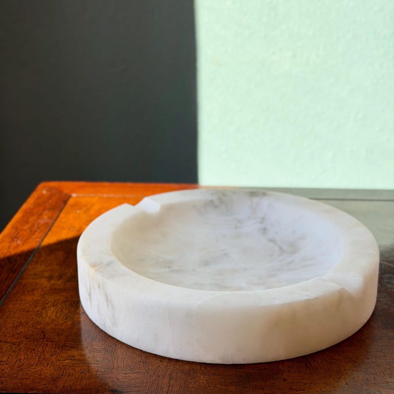 Puede incluir: Un cenicero redondo de m&aacute;rmol blanco con un solo reposacigarrillos. El cenicero tiene una superficie lisa y pulida con sutiles vetas grises. Se encuentra sobre una superficie de madera oscura, con un fondo verde y negro.