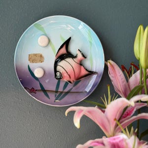 May include: A round ceramic decorative plate featuring a gradient blue and purple background with painted green seaweed. A three-dimensional pink and black angelfish is attached to the center, accompanied by two small white circular accents.