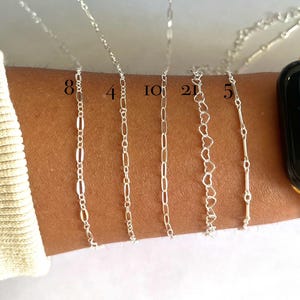 May include: A close-up of a person's wrist wearing eight different silver chains. The chains are of varying lengths and styles, including a heart-shaped chain, a box chain, and a cable chain. The chains are all silver in color and have a polished finish.