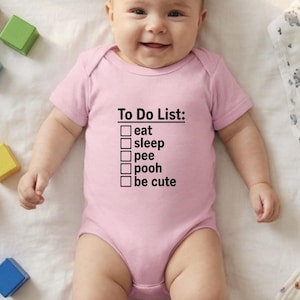 Op de afbeelding: Een lichtroze babyromper met de tekst "To Do List: eat, sleep, pee, pooh, be cute" in het zwart. De baby ligt op een witte ondergrond met kleurrijke houten blokken.