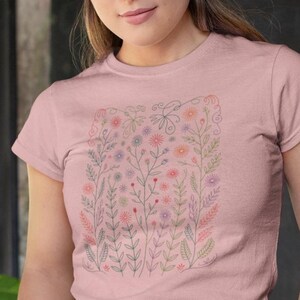 May include: A light pink t-shirt featuring a floral design. The design includes various flowers and leaves in shades of pink, purple, and green, arranged within a rectangular frame. The shirt has short sleeves and a crew neck.