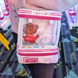 Op de afbeelding: Witte en transparante crossbodytas met roze randen en een pluchen berenspeelgoed aan de binnenkant. De tas heeft de Japanse tekst "キャッチ" en "PRIZE OUT" erop gedrukt. De tas wordt over de schouder gedragen.