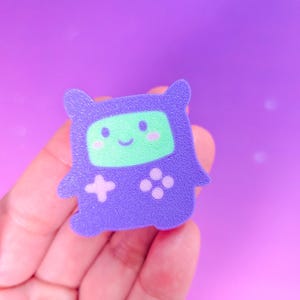 May include: A purple, cartoon-style video game character with a green screen face, pink cheeks, and a smiling mouth. The character has a cross and four-button controller design. The background is a gradient of purple and pink.