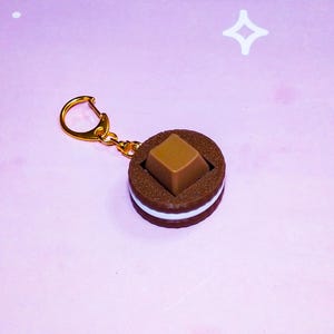 Puede incluir: Un llavero de galleta de chocolate marrón y blanco con un anillo de metal dorado. La galleta es una réplica realista de una marca popular de galletas sándwich de chocolate.