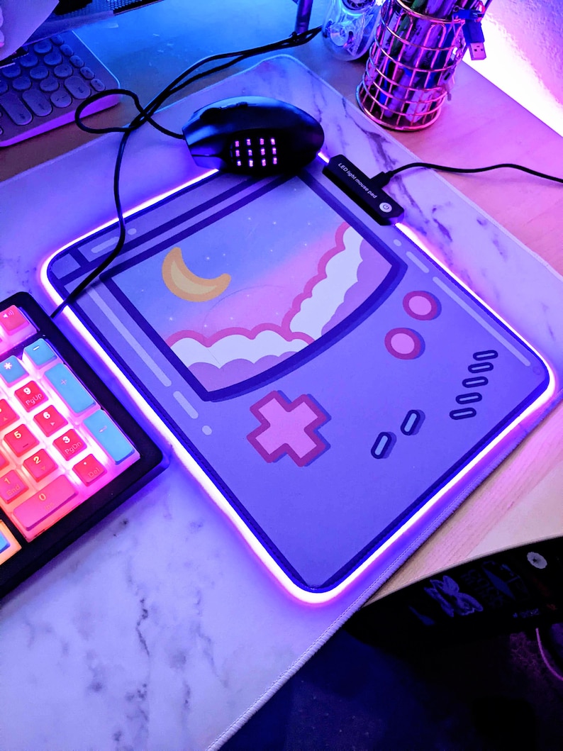 May include: A purple and pink gaming mouse mat with a retro game console design. The mouse mat has a light blue border and a glowing purple edge. The text "LED Digital Controller Pad" is visible on the mouse mat.