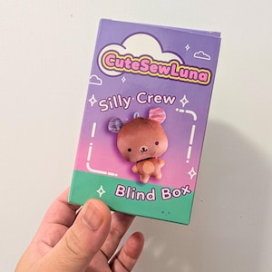 May include: A rectangular purple and green cardboard blind box featuring the text CuteSewLuna and Silly Crew. The front of the box displays a graphic of a small brown bear-like character with patterned ears and a light brown belly patch.