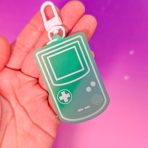 May include: A green keychain in the shape of a retro handheld game console. The keychain has a white outline and a white clip. The background is a gradient of pink and purple.