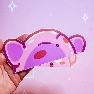 Puede incluir: Una pegatina rosa con un dise&ntilde;o de oso de dibujos animados con un cuerpo rosa claro, orejas moradas y una nariz roja. La pegatina se sostiene sobre un fondo rosa con estrellas blancas y una nube.