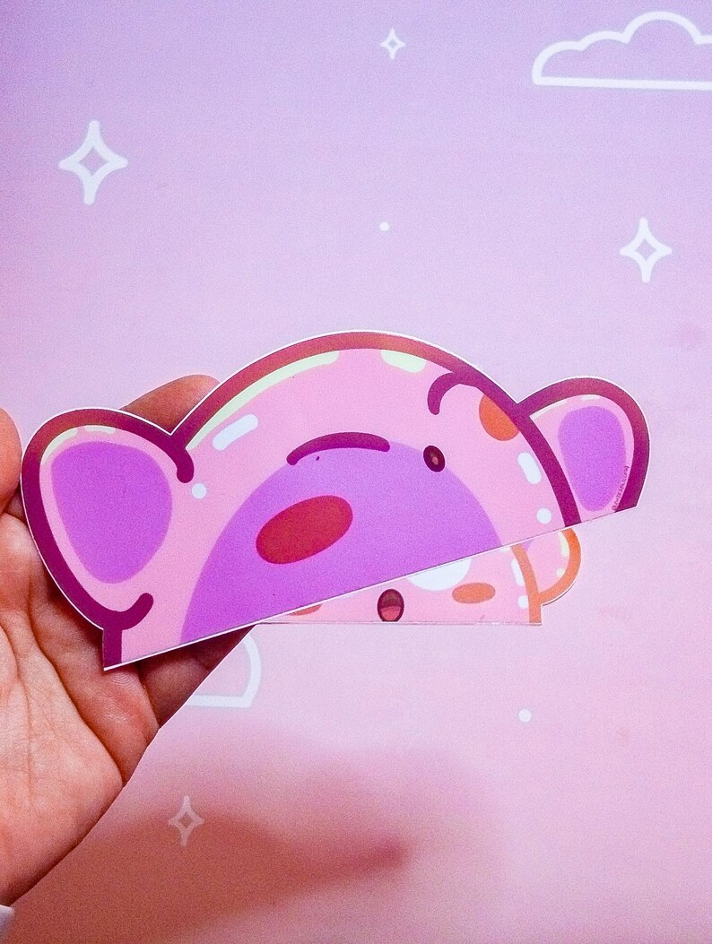 Puede incluir: Una pegatina rosa con un dise&ntilde;o de oso de dibujos animados con un cuerpo rosa claro, orejas moradas y una nariz roja. La pegatina se sostiene sobre un fondo rosa con estrellas blancas y una nube.