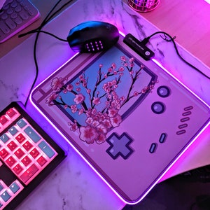 May include: A pink and purple gaming mouse mat with a pink Game Boy design and a cherry blossom tree. The mouse mat has a purple LED light around the edge.