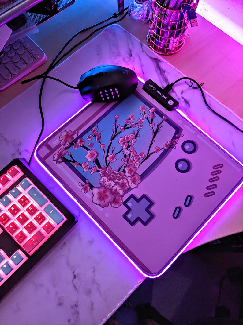 May include: A pink and purple gaming mouse mat with a pink Game Boy design and a cherry blossom tree. The mouse mat has a purple LED light around the edge.