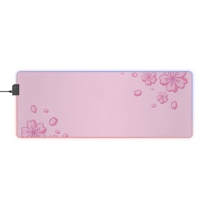 Peut inclure: Tapis de souris rectangulaire rose avec un motif floral. Le tapis présente un fond rose clair avec des illustrations de fleurs de cerisier rose foncé et une fine bordure éclairée. Un câble et un connecteur noirs sont attachés.
