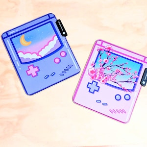 May include: Two gameboy-style phone cases, one with a pink background and a cherry blossom design, and the other with a blue background and a night sky design.