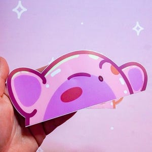 Puede incluir: Una pegatina rosa con la cara de un oso de dibujos animados con orejas grandes, una nariz roja y un peque&ntilde;o ojo negro. La pegatina se sostiene en una mano sobre un fondo rosa con estrellas blancas y una nube.