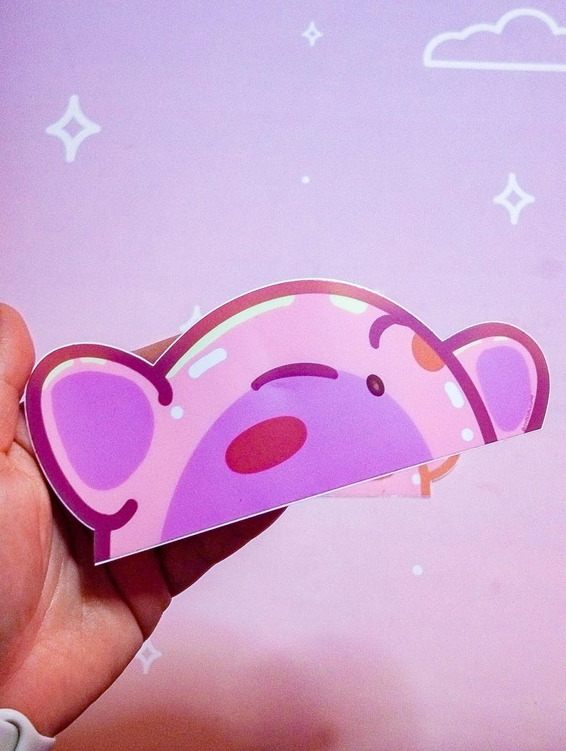 Puede incluir: Una pegatina rosa con la cara de un oso de dibujos animados con orejas grandes, una nariz roja y un peque&ntilde;o ojo negro. La pegatina se sostiene en una mano sobre un fondo rosa con estrellas blancas y una nube.