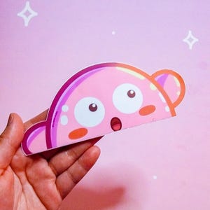 Puede incluir: Un personaje rosa de estilo caricatura con grandes ojos blancos, una expresi&oacute;n de sorpresa y peque&ntilde;as mejillas naranjas. El personaje tiene una forma curva con protuberancias en forma de oreja a cada lado. El fondo es rosa claro con estrellas blancas y una nube.