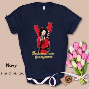 May include: Navy blue t-shirt featuring a graphic of a woman in a red outfit holding a gun, with the text "The human race is a resource" in yellow. The shirt also has the word "Navy" and size options printed on it.