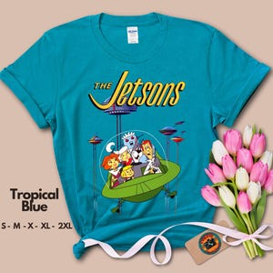 May include: A turquoise blue t-shirt with a graphic of the Jetsons family in their flying car. The text "The Jetsons" is printed in yellow above the graphic. The t-shirt is available in sizes S-M-X-XL-2XL.