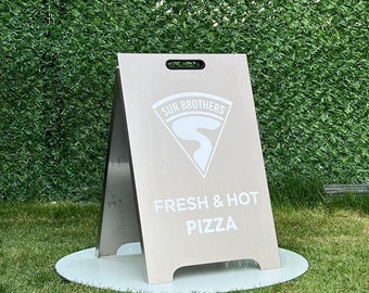 May include: A light-colored A-frame sandwich board sign featuring a white logo with the text SUN BROTHERS and the words FRESH & HOT PIZZA printed in white. The sign has a handle cutout at the top and stands on a circular white base.