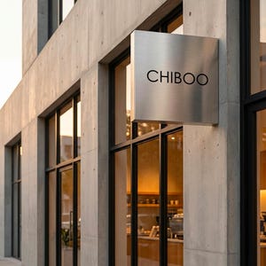 May include: Exterior view of a modern building with large windows and a concrete facade. A square, brushed metal sign with the word "CHIBOO" in black lettering hangs above the entrance. The windows reflect the interior, suggesting a cafe or shop.