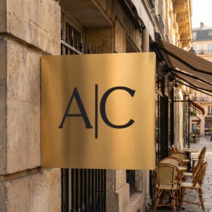 Pode incluir: Uma placa quadrada dourada com as letras pretas "A" e "C" separadas por uma linha vertical. A placa está montada em um edifício de pedra, com uma cerca de ferro forjado e assentos ao ar livre visíveis.