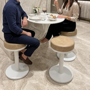 May include: A round white table with three stools featuring tan circular seats and white bases. The table is set with two wine glasses, one containing red wine and one containing white wine, a small bowl of bread, and a small vase of flowers.