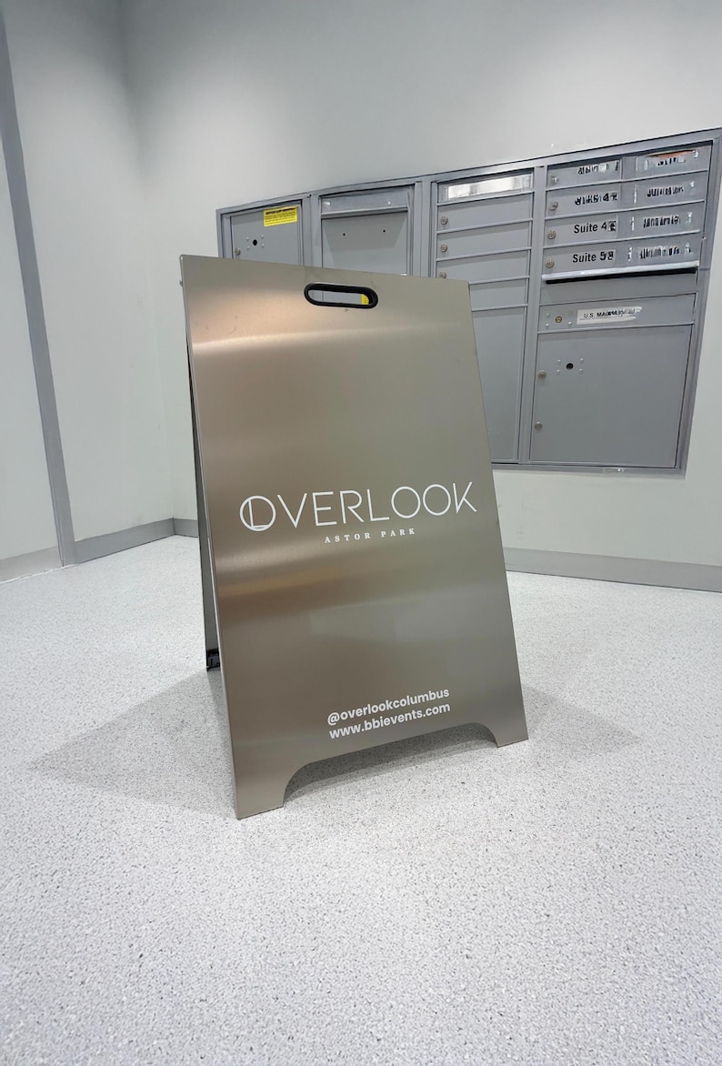 May include: A silver A-frame sign with the word "OVERLOOK" in white lettering. Below the word "OVERLOOK" is the text "ASTOR PARK". The sign also includes the website and social media handle for @overlookcolumbus and www.bbievents.com.