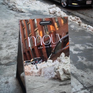 May include: A reflective silver sandwich board sign with the word "mouv" in white, along with "mind, body & soul." The sign reflects a street scene with cars and buildings. A QR code and social media handles are also visible.