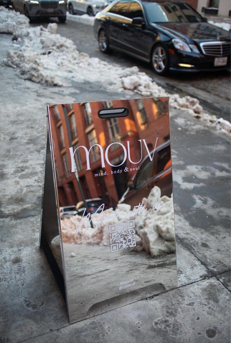 May include: A reflective silver sandwich board sign with the word "mouv" in white, along with "mind, body & soul." The sign reflects a street scene with cars and buildings. A QR code and social media handles are also visible.