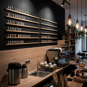 May include: A coffee shop counter featuring a black wall with wooden menu rails displaying text: HOT CHOCOLATE, MILCHKAFFEE, CAFE CREMA, ESPRESSO, LATTE MACCHIATO, CAPPUCCINO, TURKISH TEA, and TURKISH COFFEE. Includes a stainless steel sink and espresso machine.