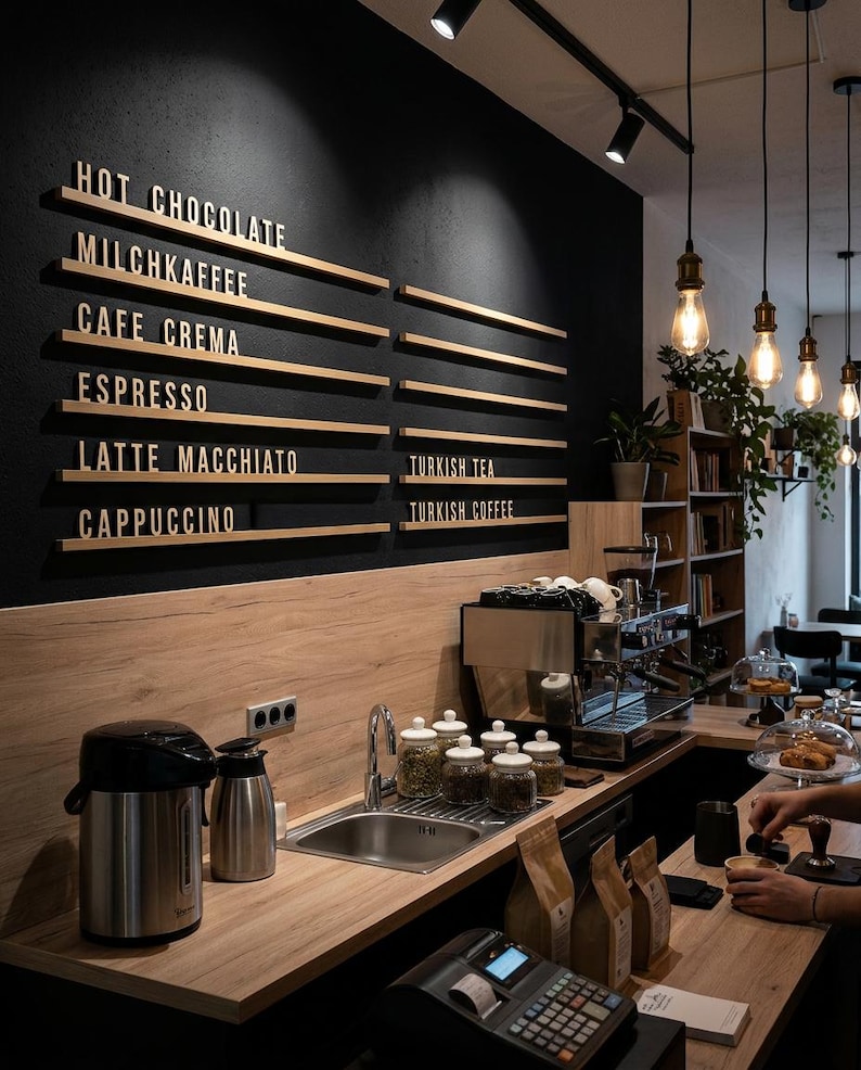 May include: A coffee shop counter featuring a black wall with wooden menu rails displaying text: HOT CHOCOLATE, MILCHKAFFEE, CAFE CREMA, ESPRESSO, LATTE MACCHIATO, CAPPUCCINO, TURKISH TEA, and TURKISH COFFEE. Includes a stainless steel sink and espresso machine.
