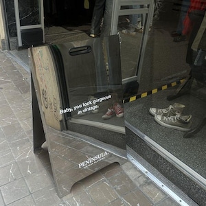 May include: A silver, reflective A-frame sidewalk sign with a handle cutout at the top. The sign features the white text "Baby, you look gorgeous in vintage." and "PEN&Iacute;NSULA VINTAGE CLOTHING" printed on the surface, reflecting the surrounding storefront.